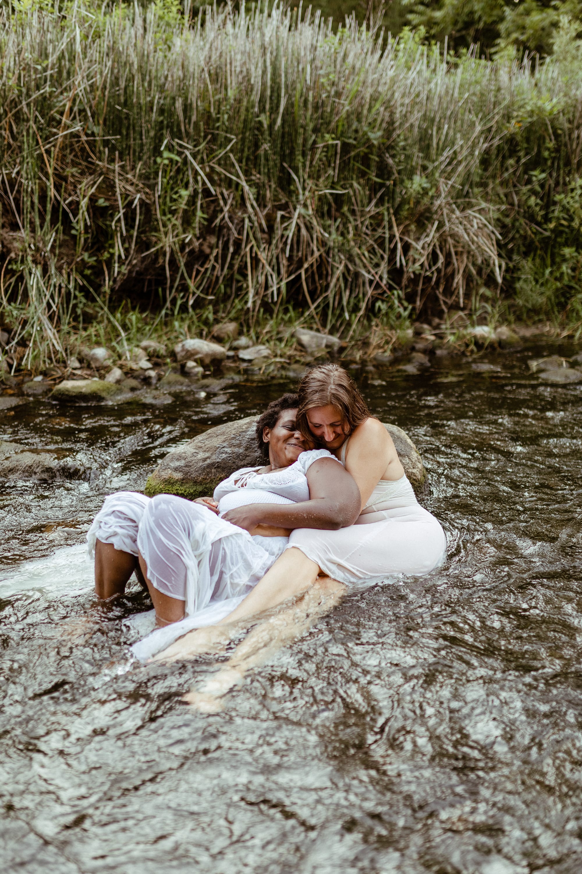 Two smiling women, one black, one white, cuddle in a bubbling creek with their backs resting on a large rock.